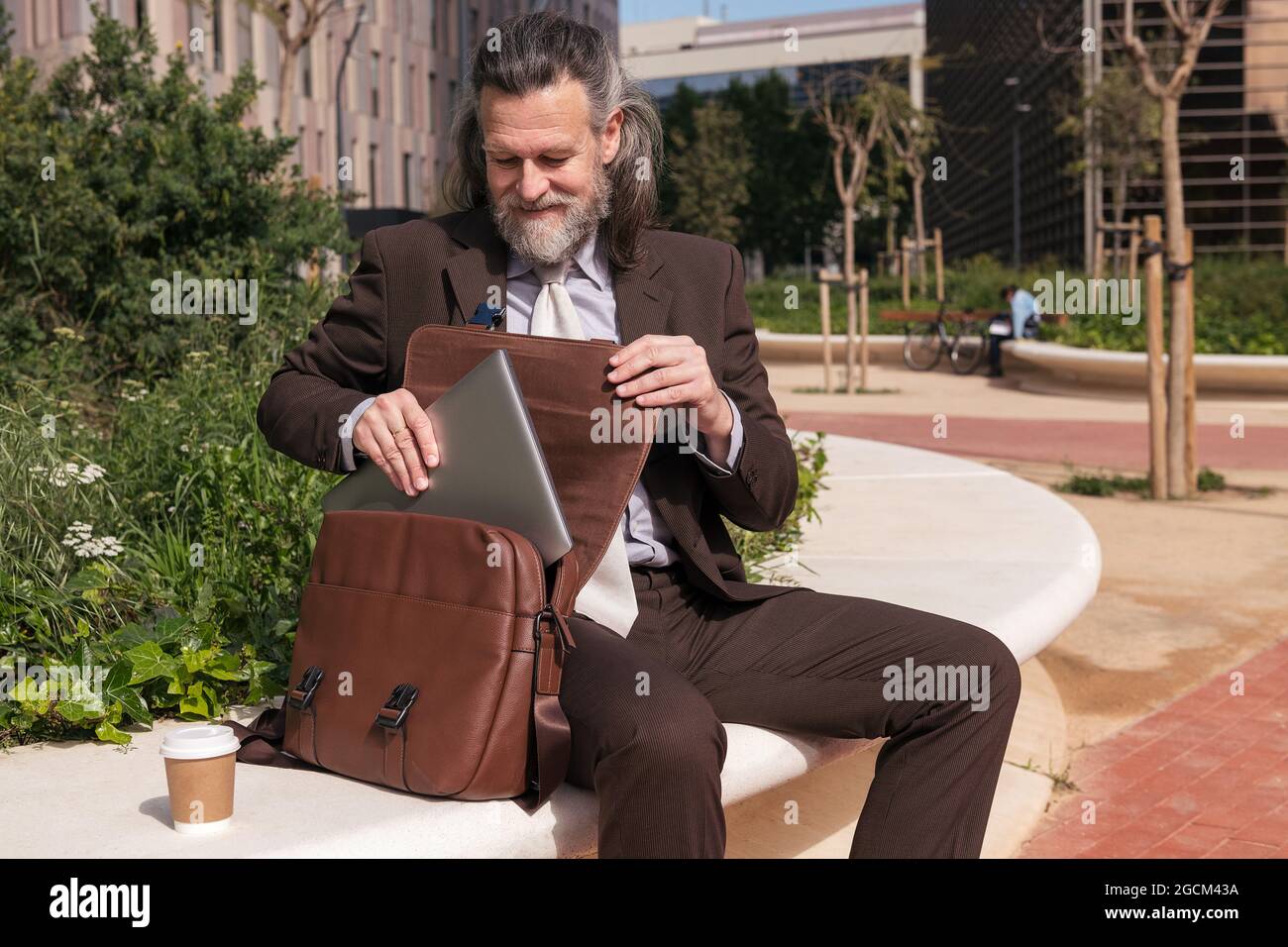 Contenido respetable de mediana edad barba hombre abogado en la ropa formal  que toma el portátil del maletín mientras se sienta con una taza de café  para llevar en urbano Fotografía de