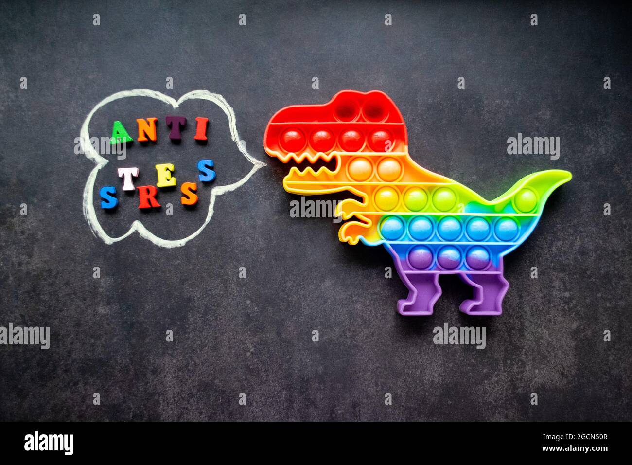 Juguete pop que dinosaurio arco iris colores sobre un fondo negro con letras multicolores y la inscripción-Anti-estrés en una burbuja de voz. Foto de stock