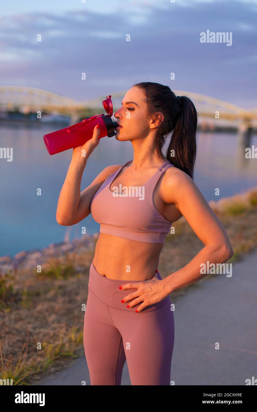 Mujer joven sedienta en ropa deportiva bebiendo refresco de botella en la orilla del río Foto de stock