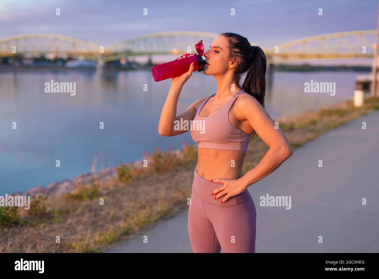 Mujer joven sedienta en ropa deportiva bebiendo refresco de botella en la orilla del río Foto de stock