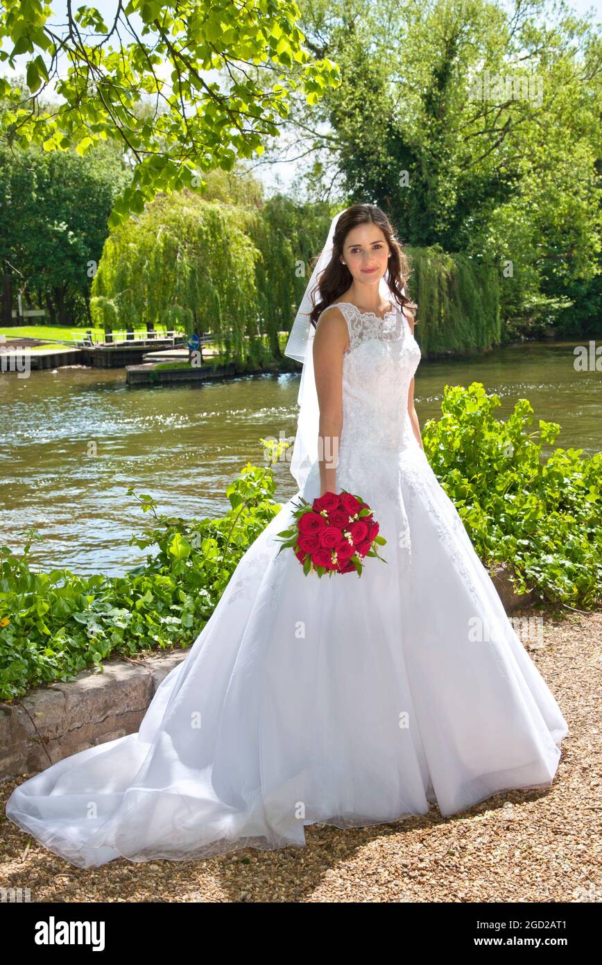 Novia en traje nupcial blanco tradicional con ramo de rosas rojas recién casado, posando en los terrenos de la iglesia junto al río con un montón de rosas rojas Foto de stock