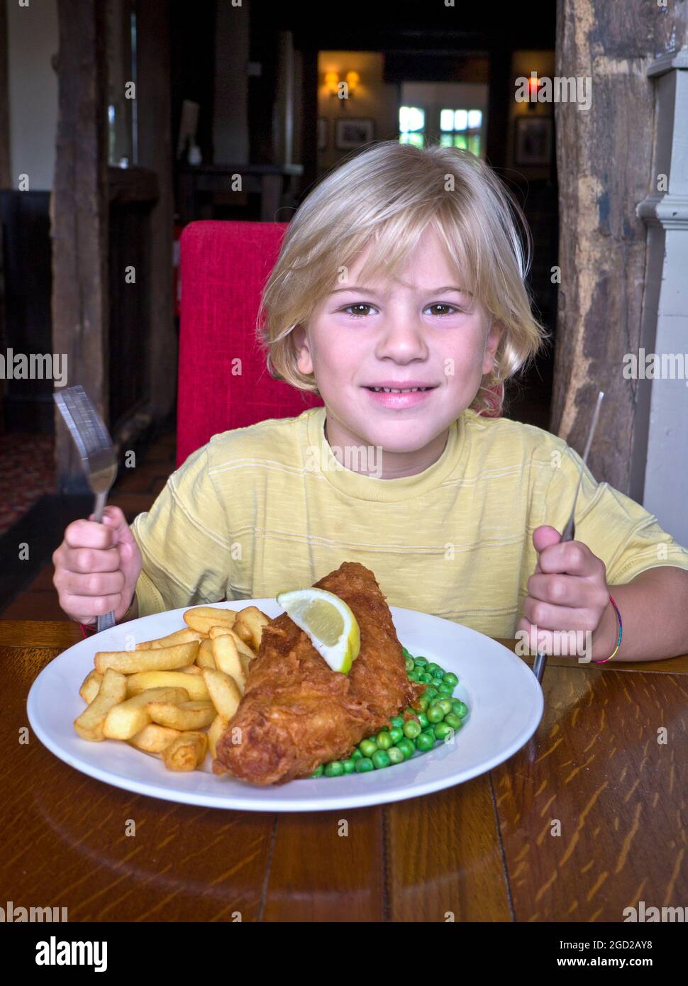 Niño de 6-8 años rubio en el interior del restaurante preparándose para comer un plato grande de pescado tradicional y patatas fritas con guisantes. Sujetar la cuchilla y la horquilla en una posición lista. Foto de stock