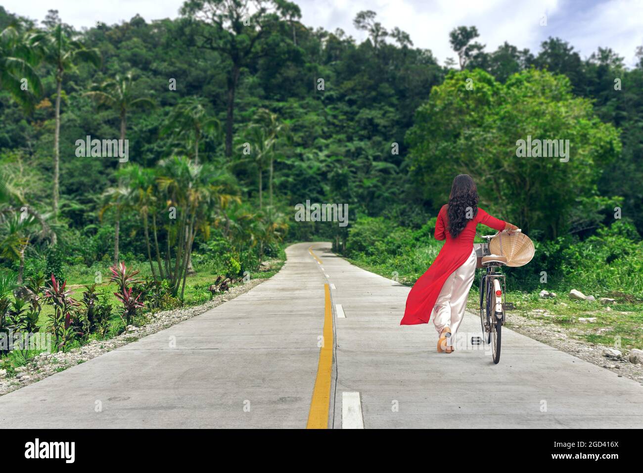 Hermosa mujer joven en el vestido tradicional vietnamita Ao Dai caminando por la carretera en la selva con bicicleta Foto de stock