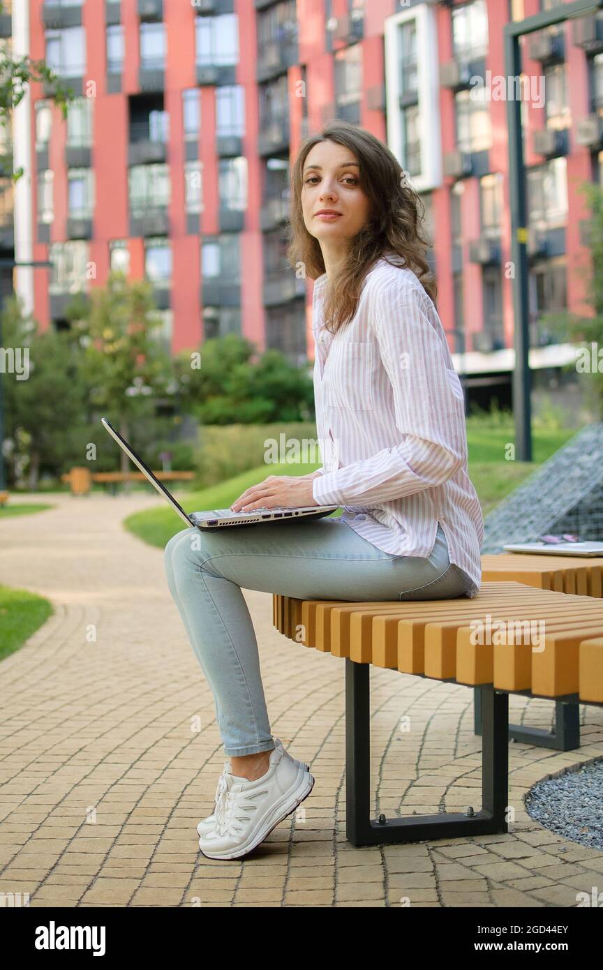 joven confiada en un traje informal está sentada en un banco al aire libre sobre un edificio moderno con un portátil gris, gerente de cuentas trabajando al libre Fotografía de