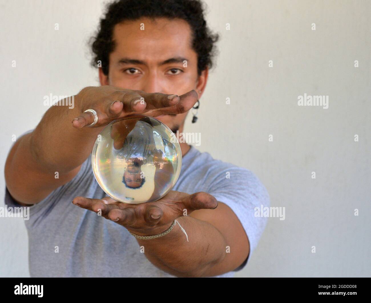 Un hombre joven guapo con uñas sucias sostiene en sus manos una bola de cristal con su propia imagen de espejo volteada verticalmente y mira al espectador. Foto de stock