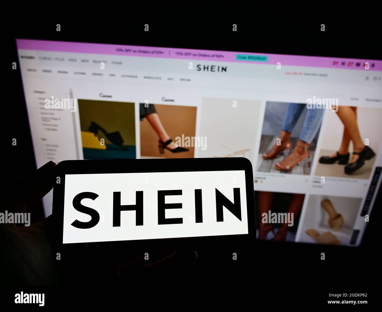 Persona que sostiene el teléfono móvil con el logotipo de la empresa china  de comercio electrónico Shein en la pantalla frente a la página web del  negocio. Enfoque en la pantalla del