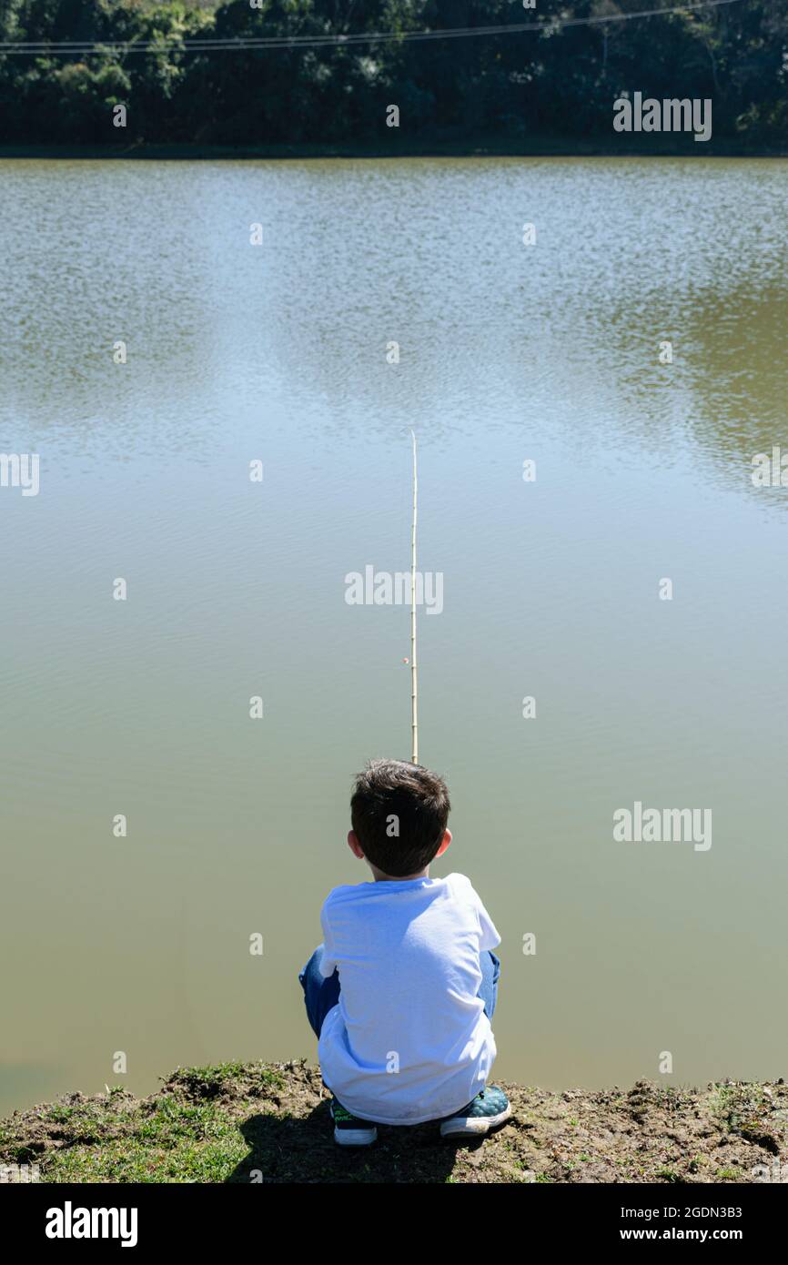 Niño brasileño de 8 años, agachado y en la espalda, pescando en una mañana soleada junto al lago (foto vertical). Foto de stock