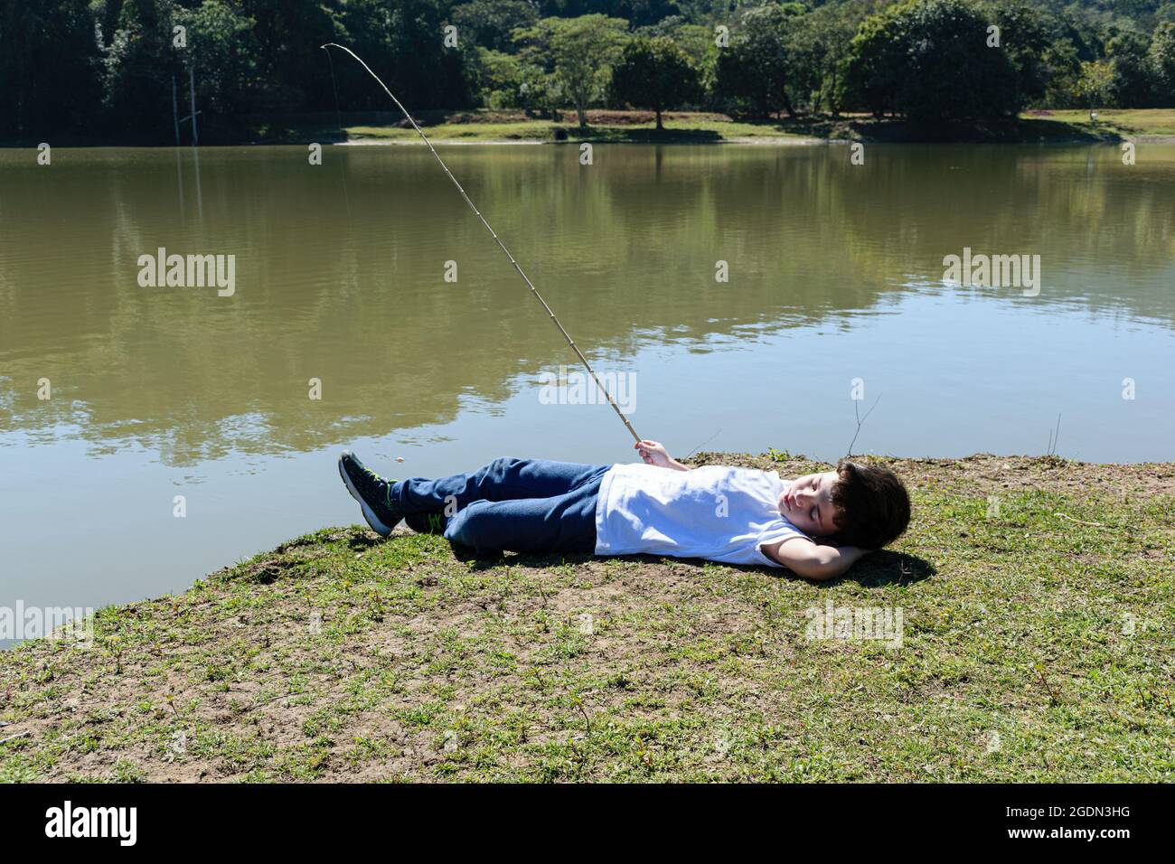 Niño brasileño de 8 años, tumbado y pescando junto al lago en una mañana soleada. Foto de stock