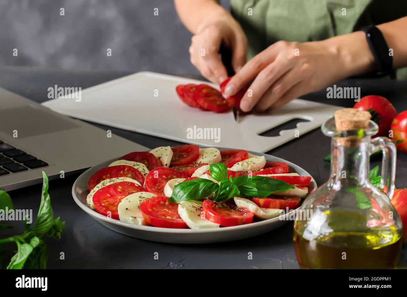 Mujer joven viendo clase magistral culinaria en línea y aprendiendo a cocinar ensalada tradicional italiana de capreses con tomates frescos, queso mozzarella y Foto de stock