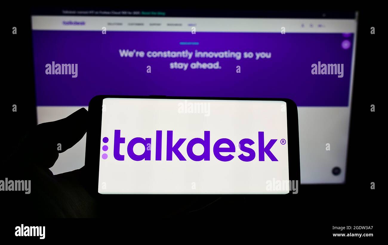 Persona que sostiene el teléfono móvil con el logotipo de la compañía de centros de contacto de EE.UU. Talkdesk Inc. En la pantalla frente a la página web de la empresa. Enfoque en la pantalla del teléfono. Foto de stock