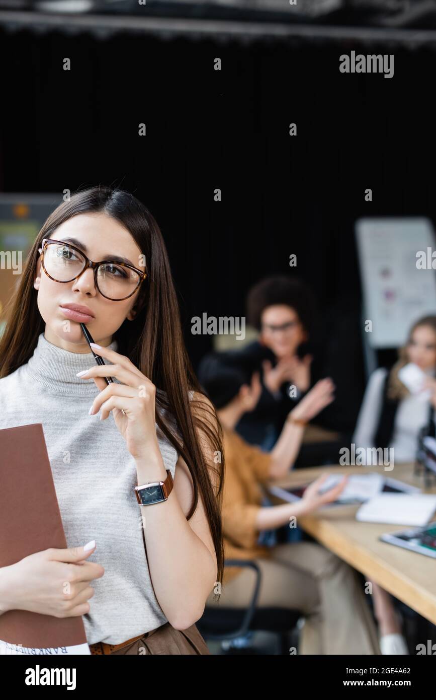 mujer de negocios con carpeta de papel y lápiz pensando cerca de los gerentes que trabajan sobre fondo borroso Foto de stock