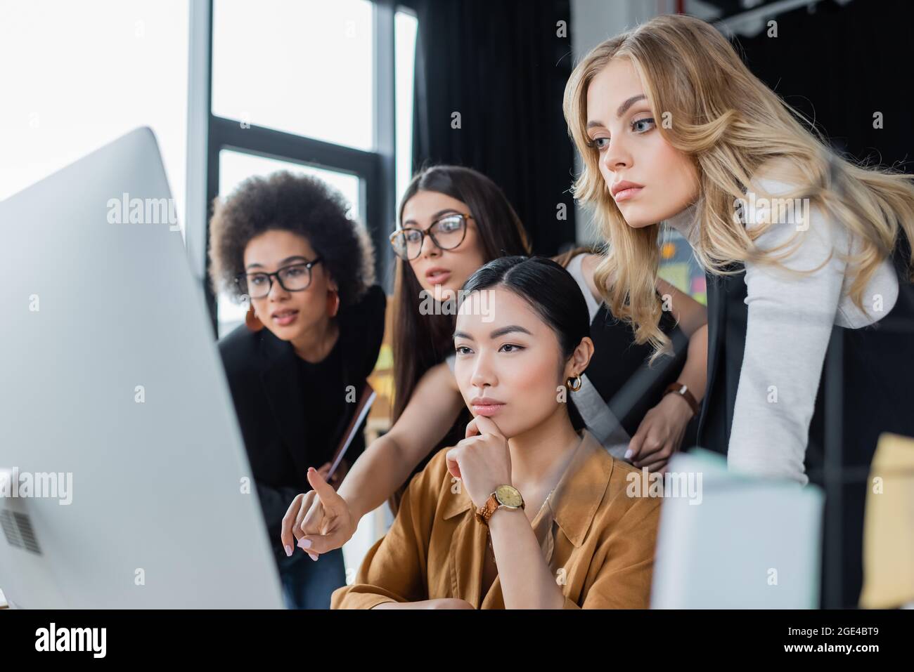 empresaria emocionado apuntando al monitor de computadora borrosa cerca de colegas multiétnicos reflexivos Foto de stock