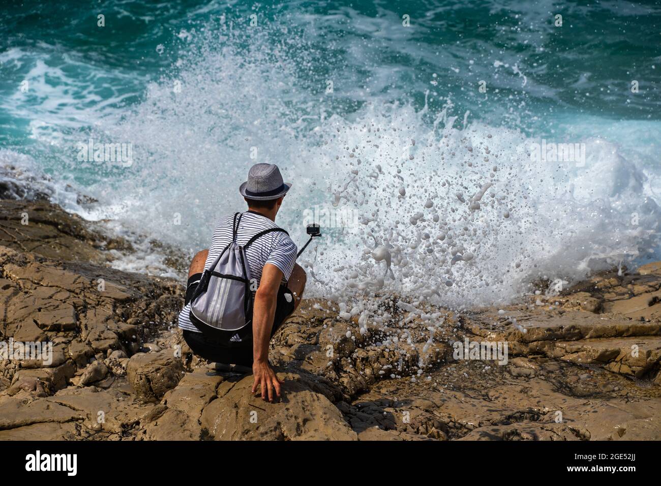 Joven tomando fotos de olas rebotando sobre rocas resbaladizas con una cámara de acción en Primosten, Croacia Foto de stock