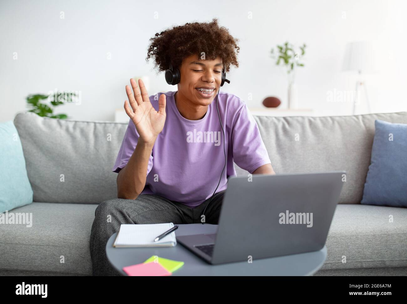 Feliz adolescente negro en auriculares saludando a su amigo o profesor en línea, agitando en la cámara desde casa. Estudiante adolescente afroamericano positivo que nos tiene Foto de stock