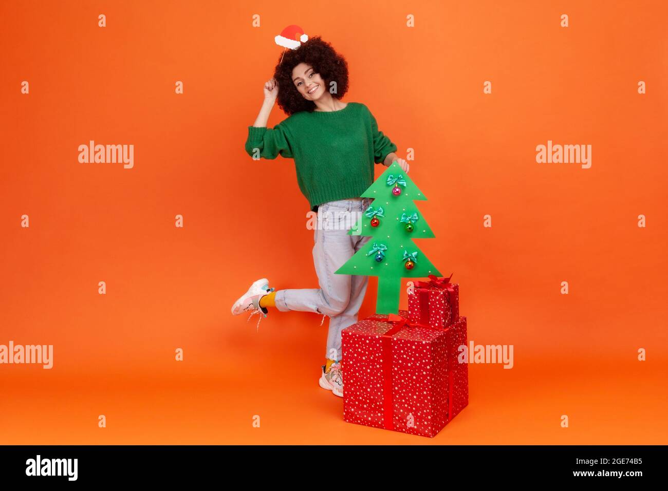 Longitud completa de mujer con pelo rizado vestido verde sport estilo suéter que sostiene la máscara en el palo con sombrero de santa claus cerca del árbol de Navidad y cajas de Longitud completa de mujer con pelo rizado vestido verde sport estilo suéter que sostiene la máscara en el palo con sombrero de santa claus cerca del árbol de Navidad y cajas de