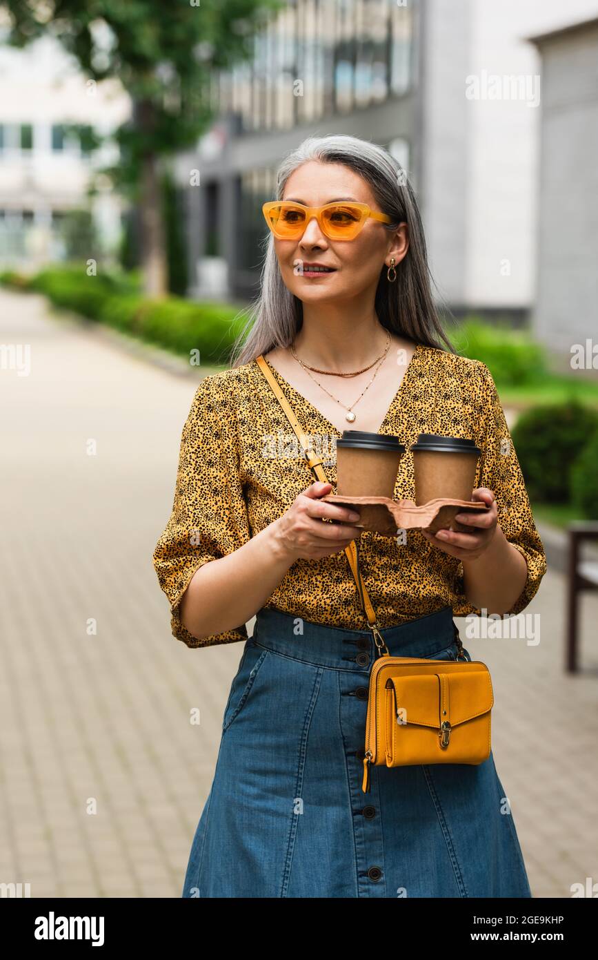 mujer madura con blusa estampada y gafas de sol amarillas de pie con café  para ir a la calle Fotografía de stock - Alamy