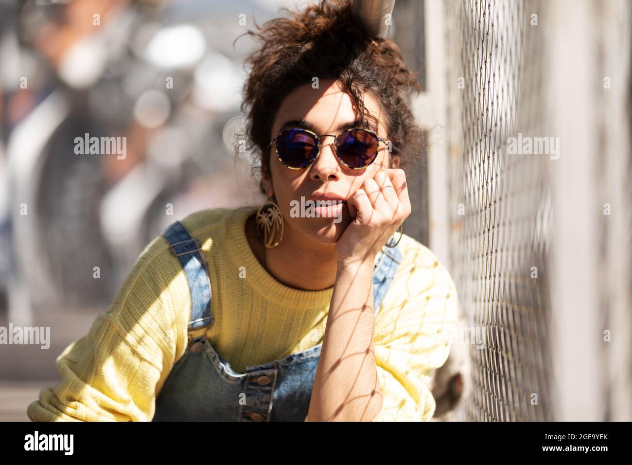 Moderna mujer hispana milenaria con cabello rizado vestido con sudadera  amarilla con monos vaqueros y gafas de sol y pendientes de moda apoyados  Fotografía de stock - Alamy
