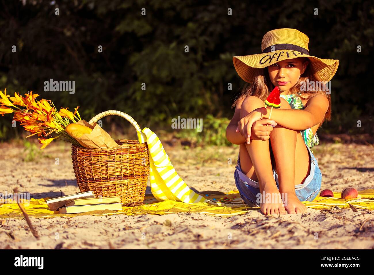 Hermosa adolescente un sombrero con una lollipop sandía se sienta en una playa de arena en el soleado día de verano. Niña linda en un picnic. Cesta mimbre, bagu Fotografía