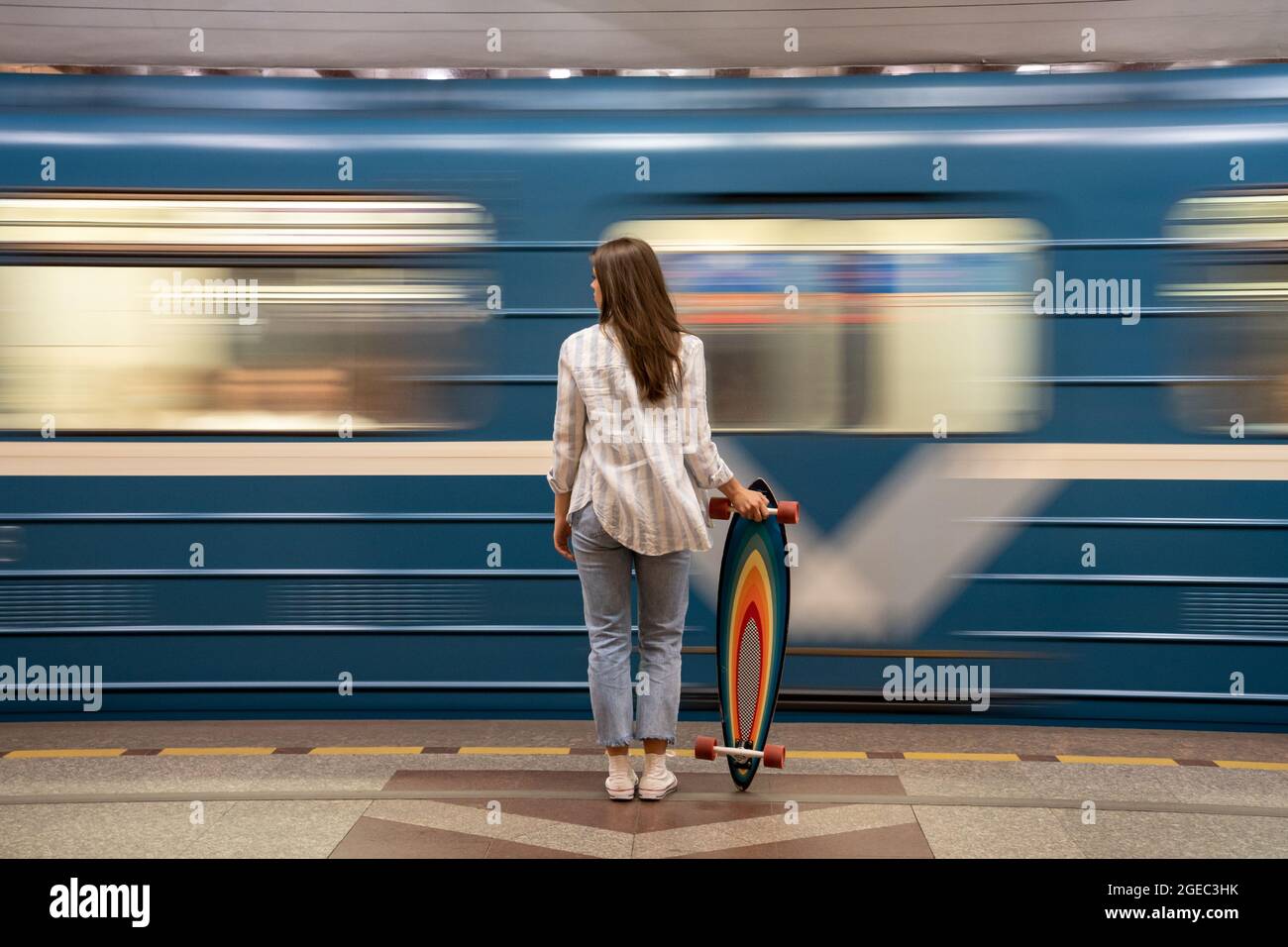Chica con longboard de pie en plataforma subterránea con un tren azul borroso en movimiento sobre el fondo Foto de stock