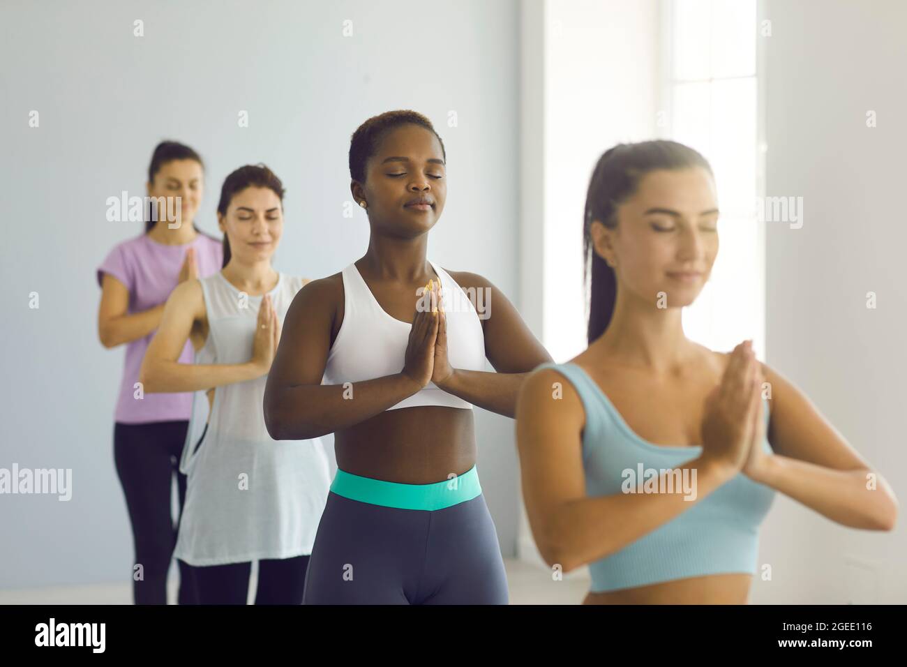 Mujer joven afro americana relajada de pie con brazos doblados en el gesto namaste en la clase de yoga en grupo. Foto de stock