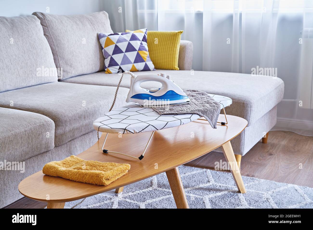 Toallas lavadas en una tabla de planchar y una plancha en una pequeña sala  de estar con sofá, tiempo de rutina del hogar