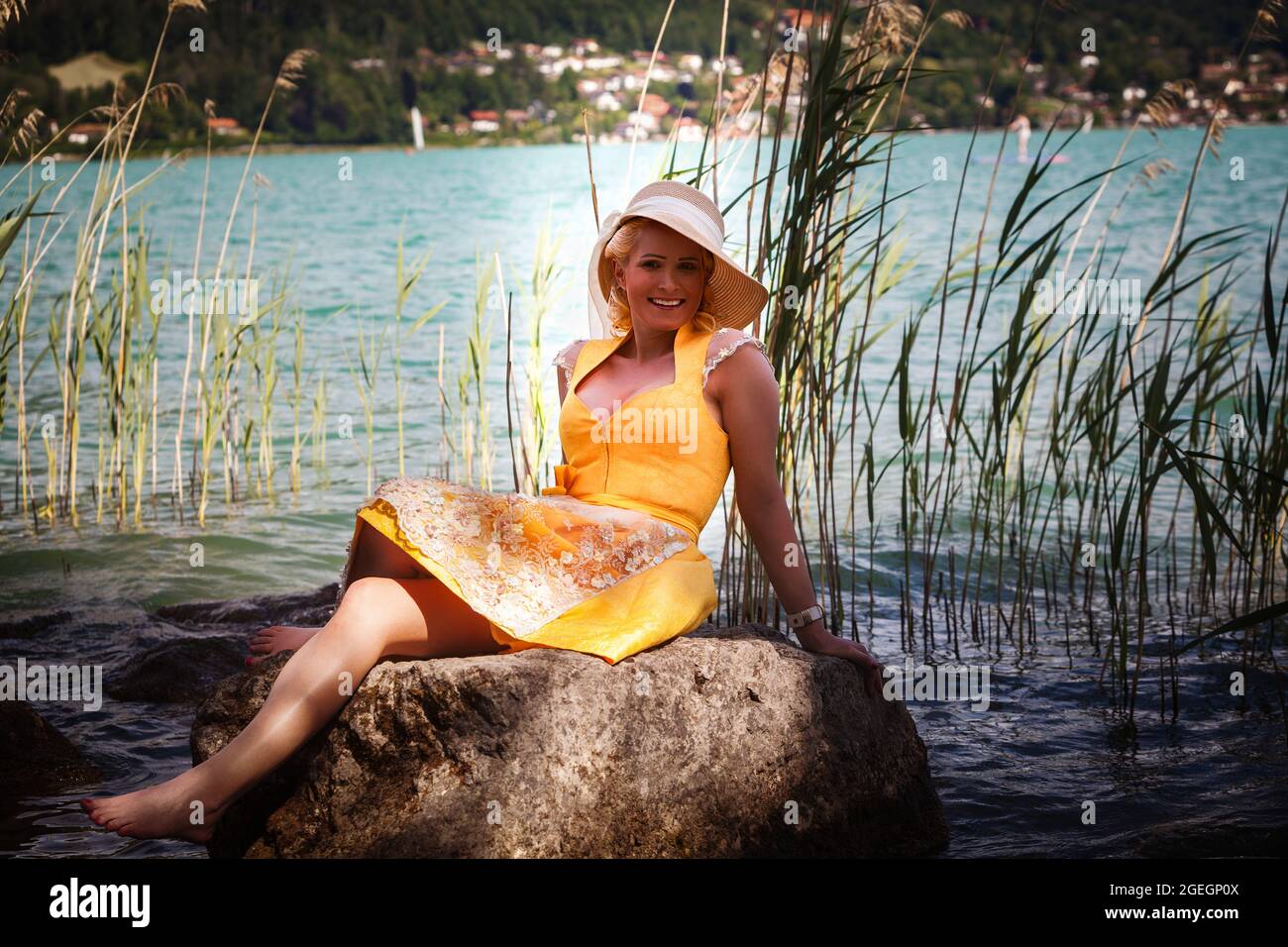 Mujer elegante con dirndl y sombrero de sol se sienta sonriendo en una roca en un lago Foto de stock