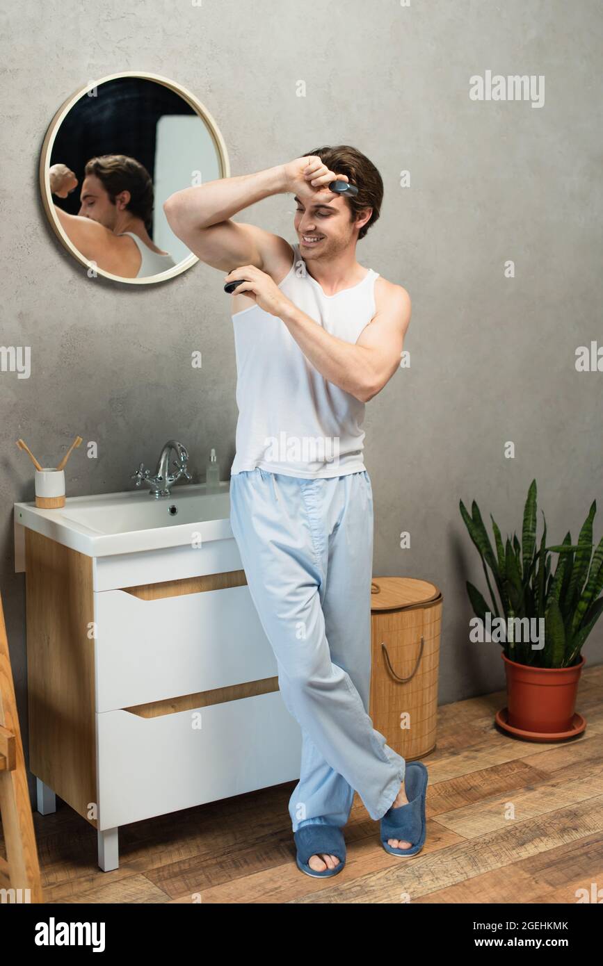 hombre sonriente camiseta de tirantes blanca y pantalones de pijama azules que aplican desodorante en la axila Fotografía de stock - Alamy