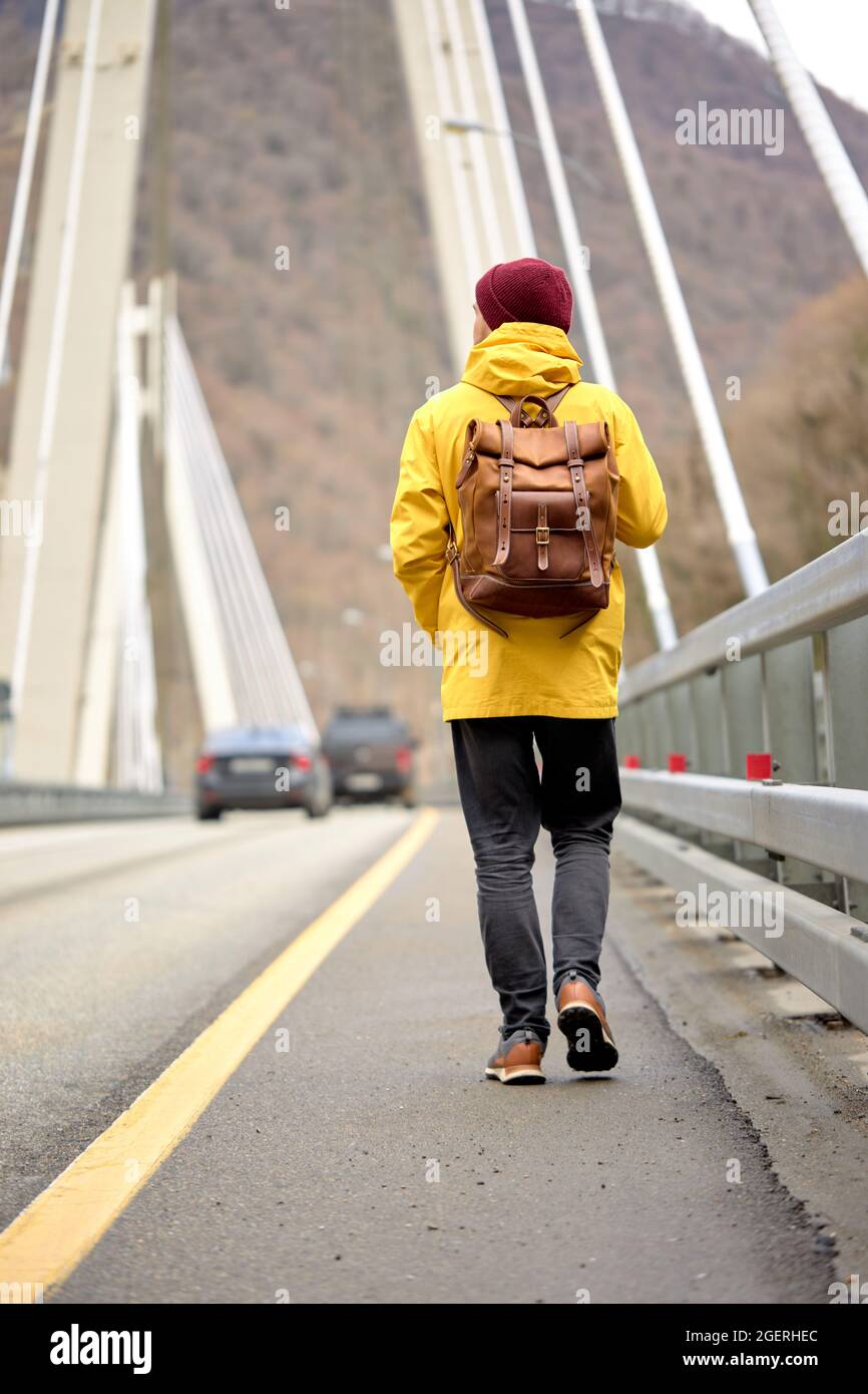 Hombre de abrigo amarillo caminando solo por un camino peatonal en el  puente de un parque urbano. Estilo de vida saludable, viajes, aventura,  concepto de senderismo. Vista trasera Fotografía de stock -