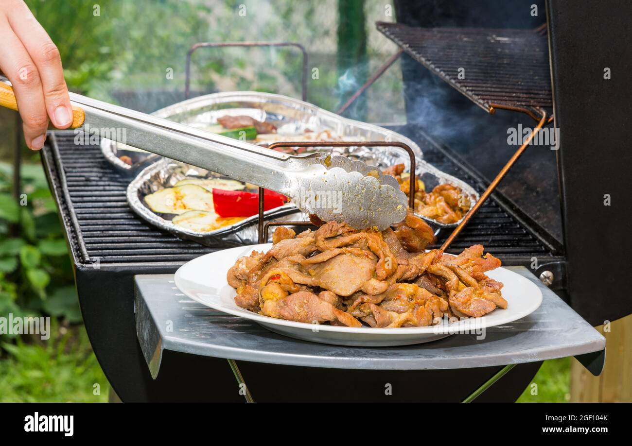 Parrilla de carne de cerdo caliente y rodajas de verduras en parrilla de  carbón en la barbacoa del jardín. Detalle de las pinzas metálicas de  sujeción de las manos humanas. Barbacoa en