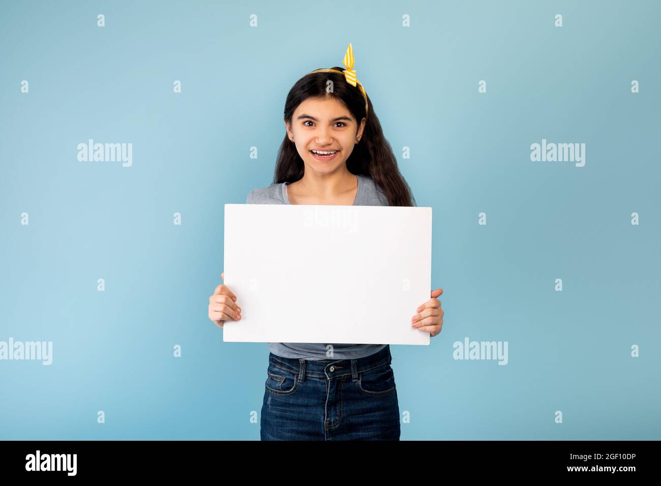 Increíble promoción. Feliz niña adolescente india sosteniendo pancarta blanca en blanco con espacio de copia sobre fondo azul del estudio, burlándose Foto de stock