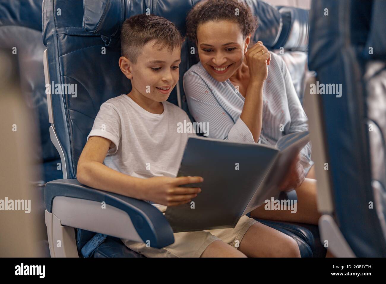 Niño, niño a bordo sosteniendo un libro y mirando a través de él junto con su mamá durante el vuelo. Tripulación de cabina que proporciona servicio a la familia en avión Foto de stock
