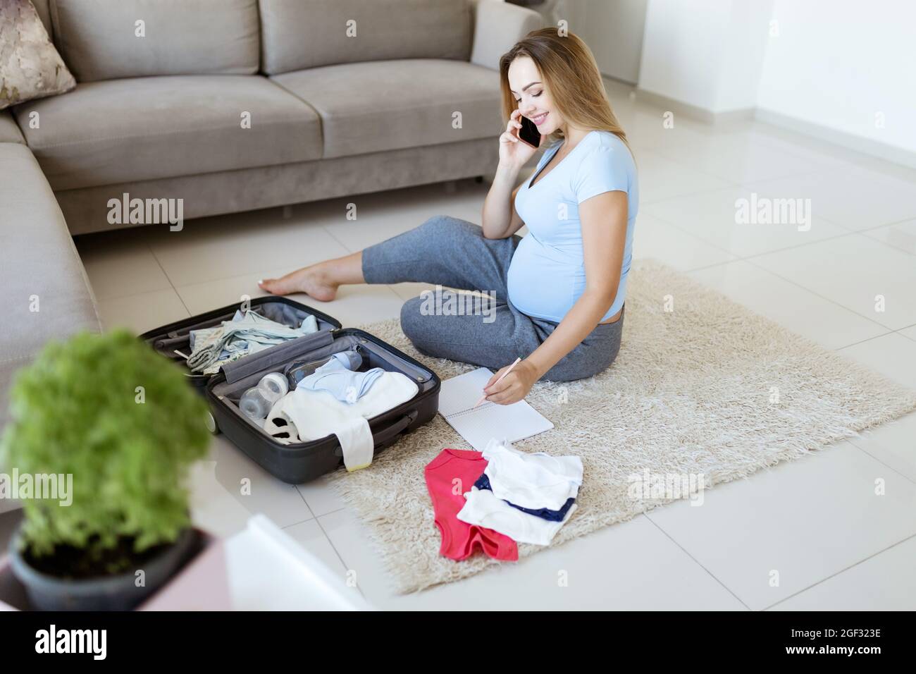 Mujer embarazada en camiseta azul, lista de verificación, teléfono de habla  y preparación de cosas para el hospital de maternidad en casa. Feliz  embarazo empacando ropa de bebé en la maleta Fotografía