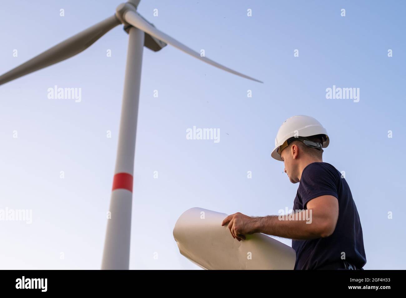 Un ingeniero de aerogeneradores que tiene un proyecto de papel y que mira el molino de viento en el campo. Generación ecológica de electricidad y energía renovable. Foto de stock