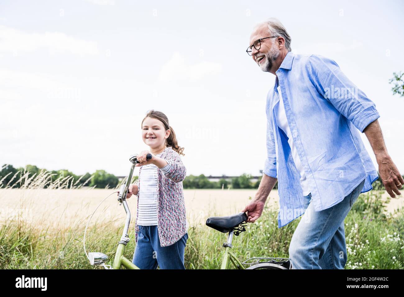 Sonriente abuelo caminando con bicicleta y nieta en el prado Foto de stock