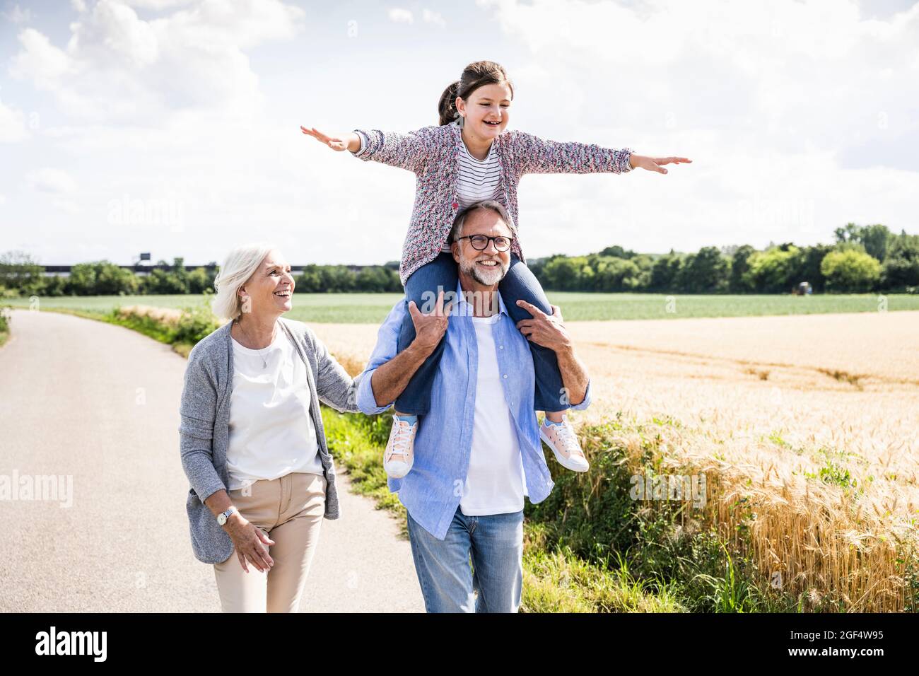 Abuelo llevando alegre nieta en el hombro mientras camina por mujer en la carretera Foto de stock