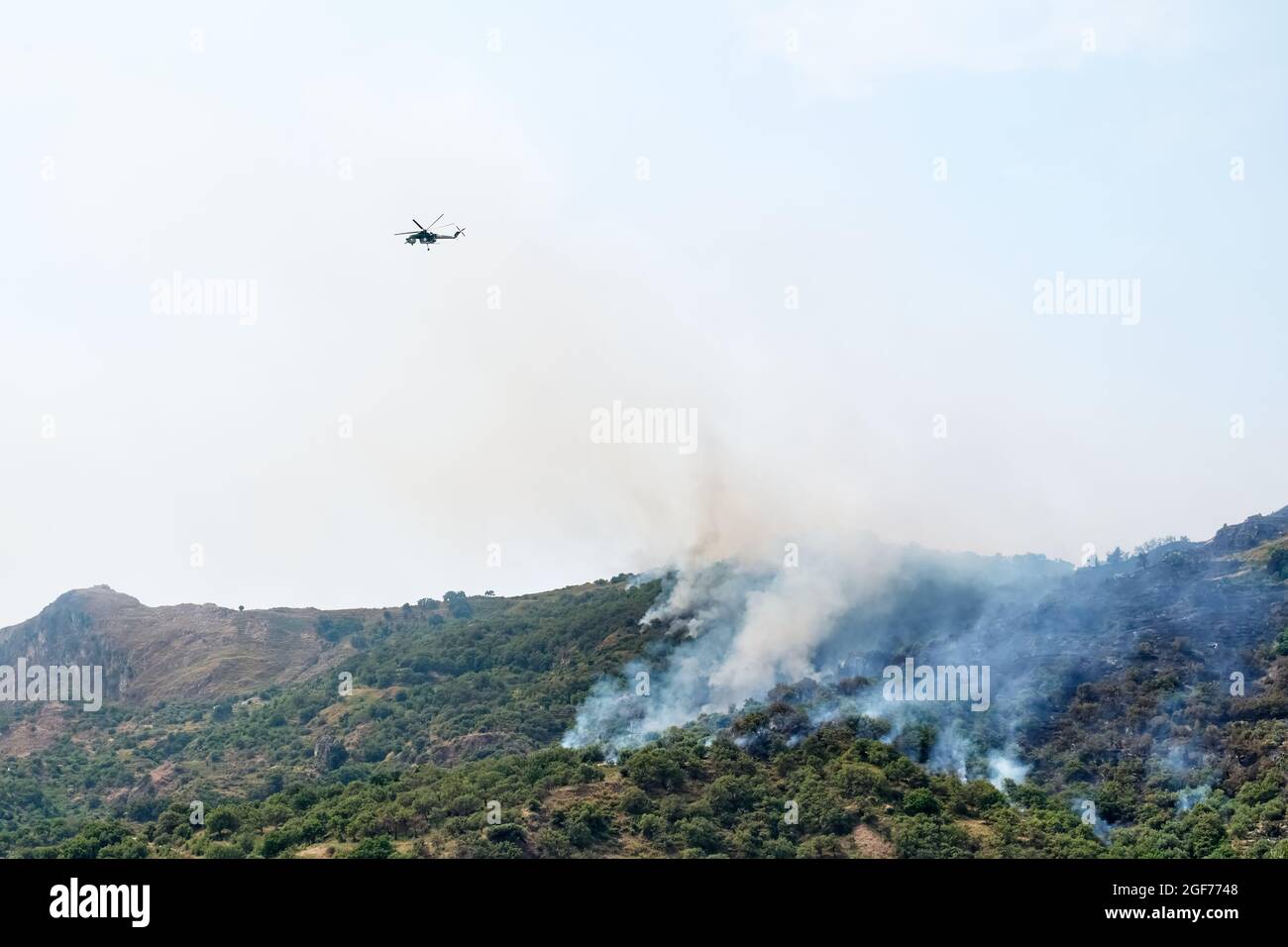 Fuego helicóptero que cae agua de un cubo en un fuego de bosque en las montañas. Los incendios forestales son un desastre ambiental. Deforestación. Foto de stock