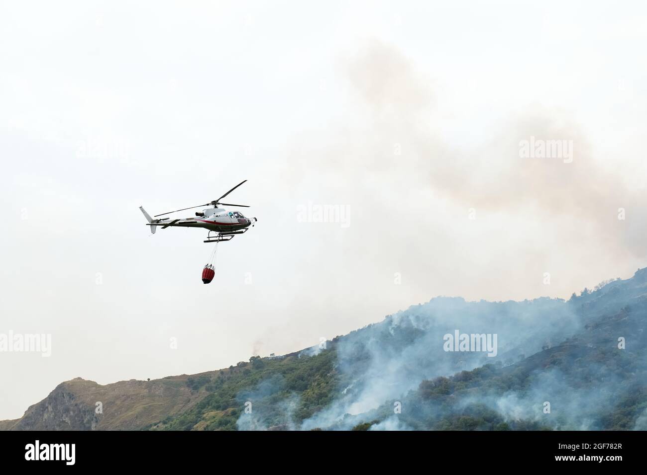 Fuego helicóptero que cae agua de un cubo en un fuego de bosque en las montañas. Los incendios forestales son un desastre ambiental. Deforestación. Foto de stock