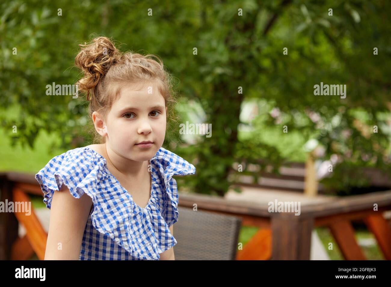 Adorable chica con ropa de cuadros mirando la cámara mientras se relaja en el patio el día de fin de semana de verano Foto de stock