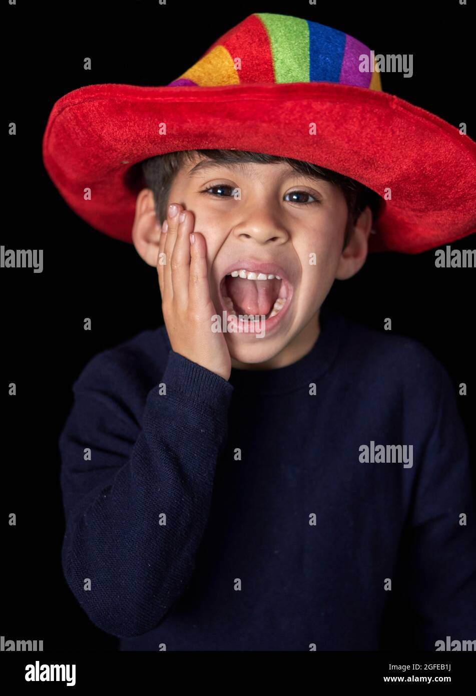 niño latino con sombrero rojo haciendo caras y apoyando su mano en su cara gritando. fondo vertical, negro Foto de stock