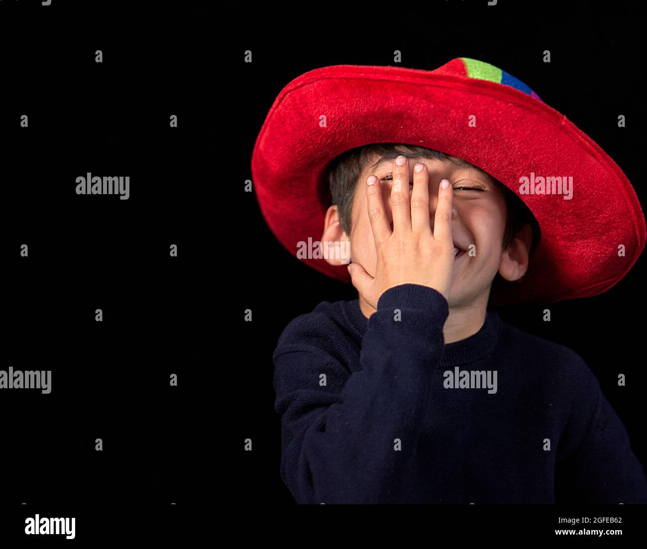 Niño latino con un sombrero rojo sonriendo y cubriendo su cara con su mano. Espacio de copia y fondo negro Foto de stock