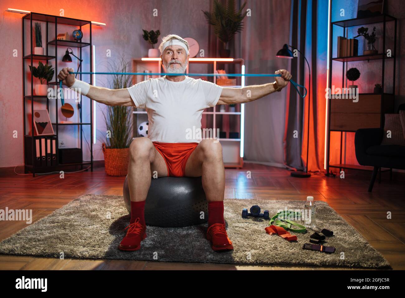 Hombre alto activo en los brazos de entrenamiento de ropa deportiva con  banda de resistencia mientras está sentado en el balón de equilibrio.  Persona mayor que pasa tiempo libre en casa para