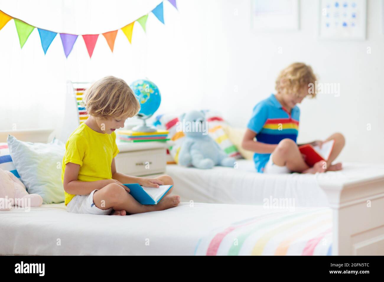 Niño leyendo libro en la cama. Habitación para dos niños. Los niños  pequeños leen libros en una cama blanca con ropa de cama colorida.  Guardería infantil. Niños chicos hermanos jugando Fotografía de