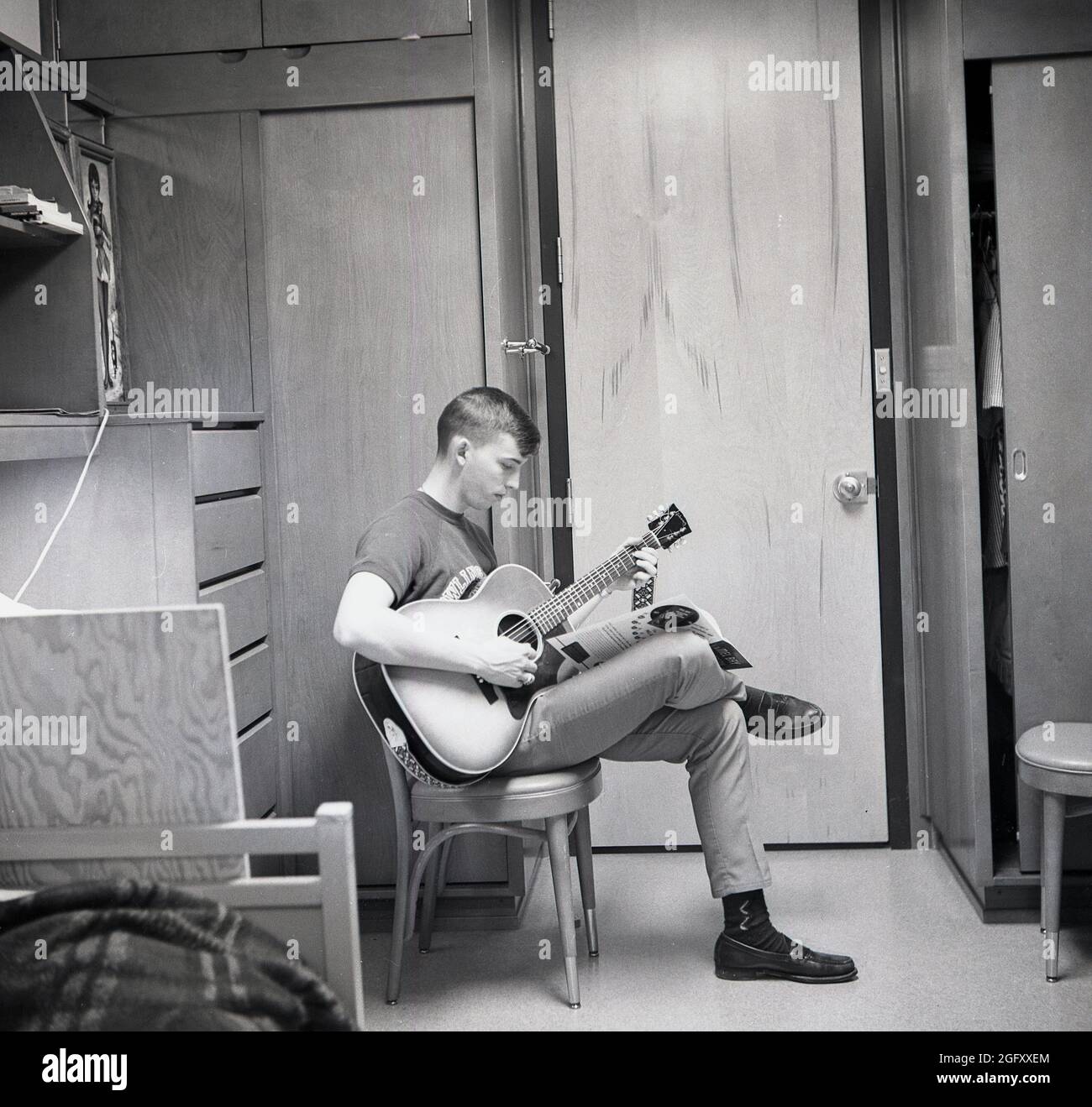 1959, histórico, un estudiante universitario masculino sentado en su  dormitorio, con un folleto en su regazo aprendiendo a tocar la guitarra  acústica, EE.UU.. Una guitarra acústica es una guitarra tradicional, no  amplificada,