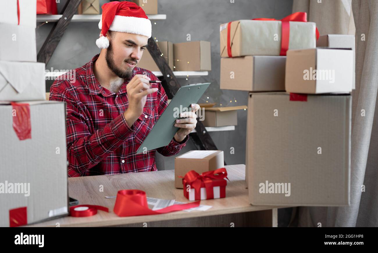 Retrato de un tipo barbudo Santa Claus en un sombrero trabaja en un almacén de una tienda en línea prepara las mercancías en cajas para el envío para la Navidad. Año Nuevo 20 Foto de stock