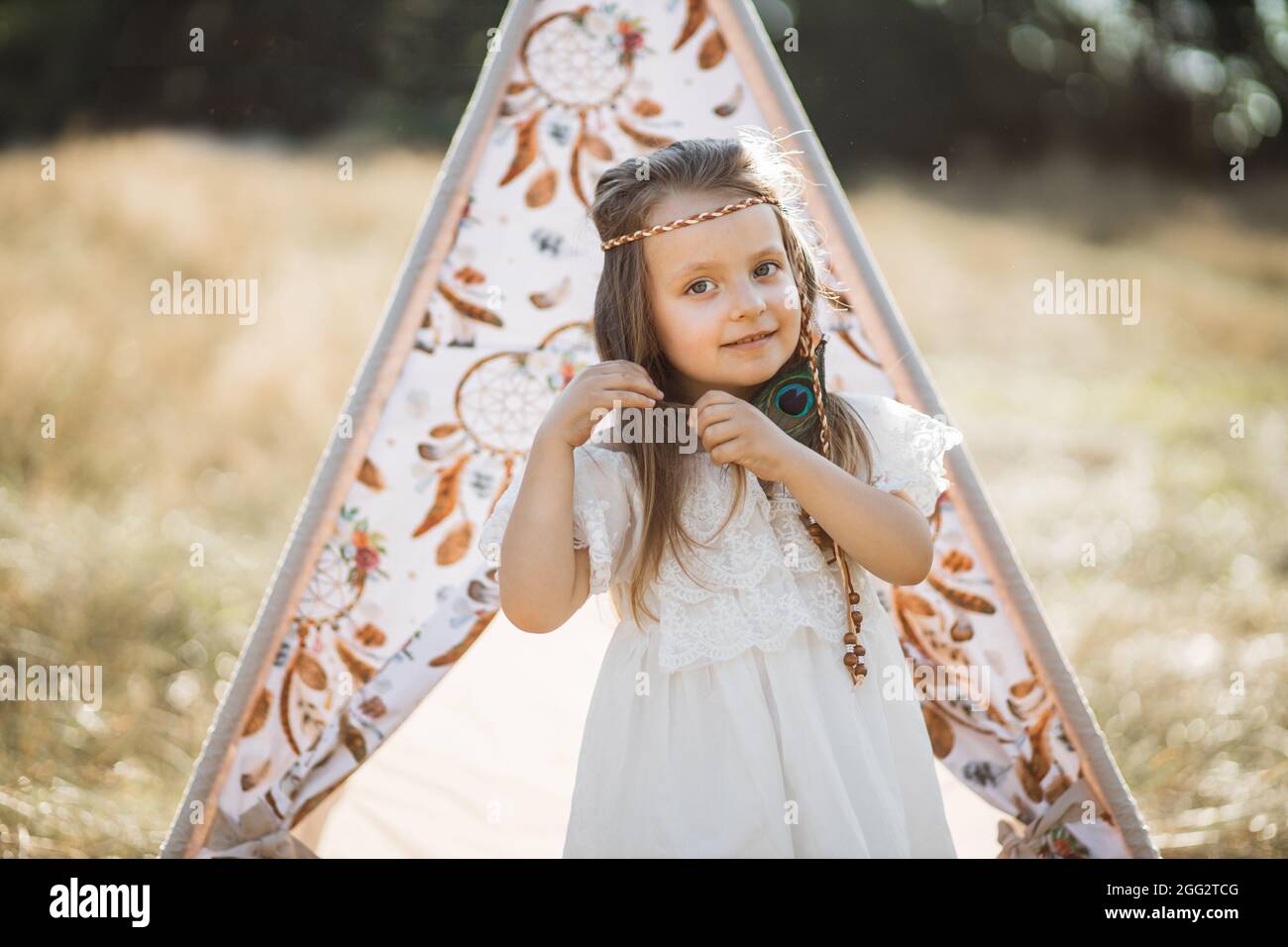 Primeros planos al aire libre de encantadora niña adorable, vestido blanco  y vestido de plumas, posando a la cámara cerca de elegante tienda de tetis  en hermoso campo de verano salvaje Fotografía