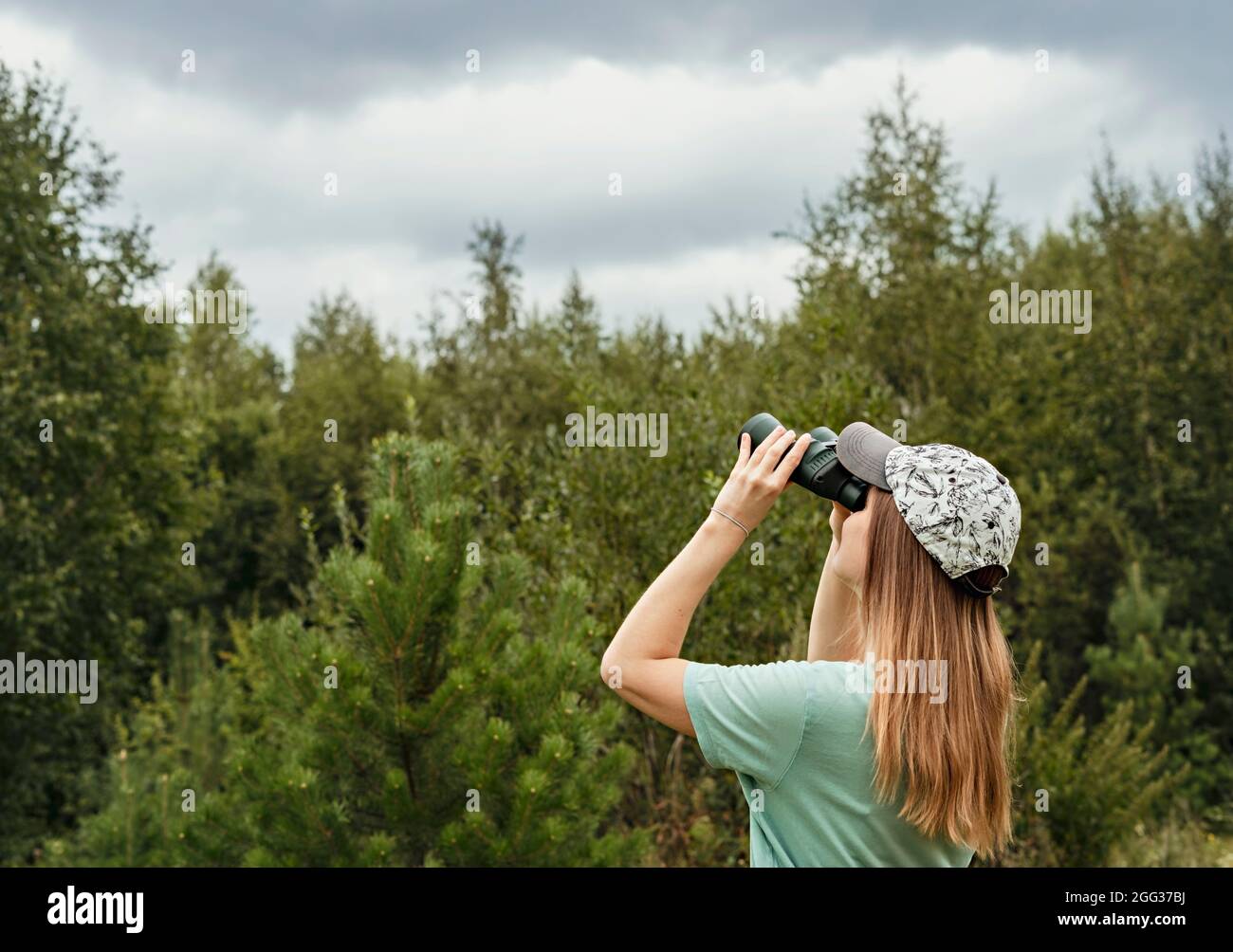 Joven mujer rubia observador de aves en gorra y azul mirando a través de binoculares en cielo nublado en verano bosque ornitológico investigación Birdwatching, zoolog Foto de stock
