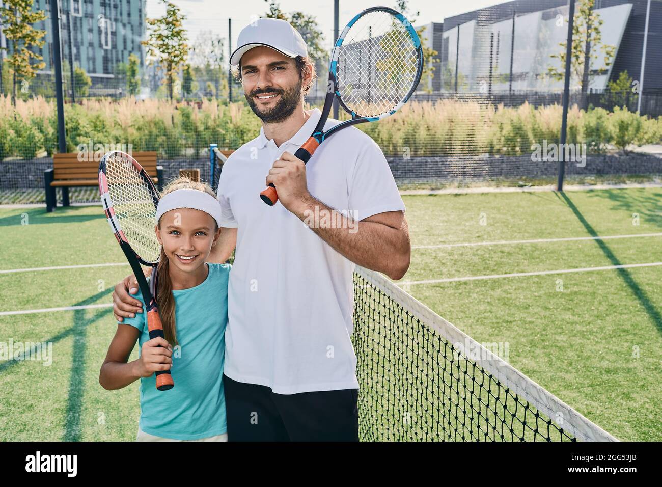 Retrato del jugador de tenis padre e hija pequeña en ropa deportiva con raquetas en las manos en la cancha de tenis al aire libre cerca de la red. familia deportiva jugando tenni Foto de stock
