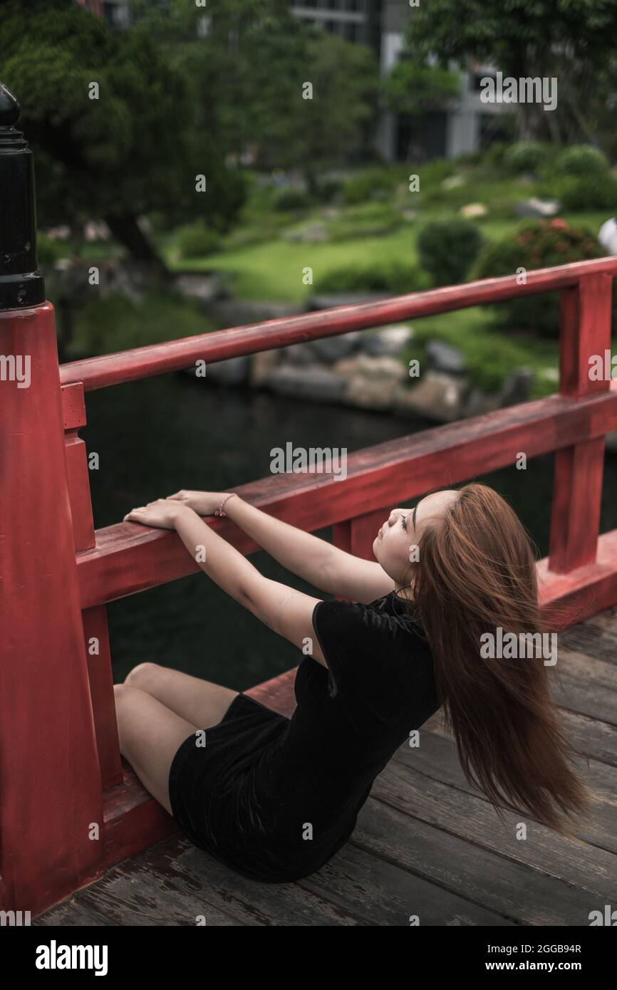 Retrato de una hermosa mujer asiática joven con un vestido negro informal sentado en el parque en el puente con pasamanos. Encantadora chica vietnamita Foto de stock