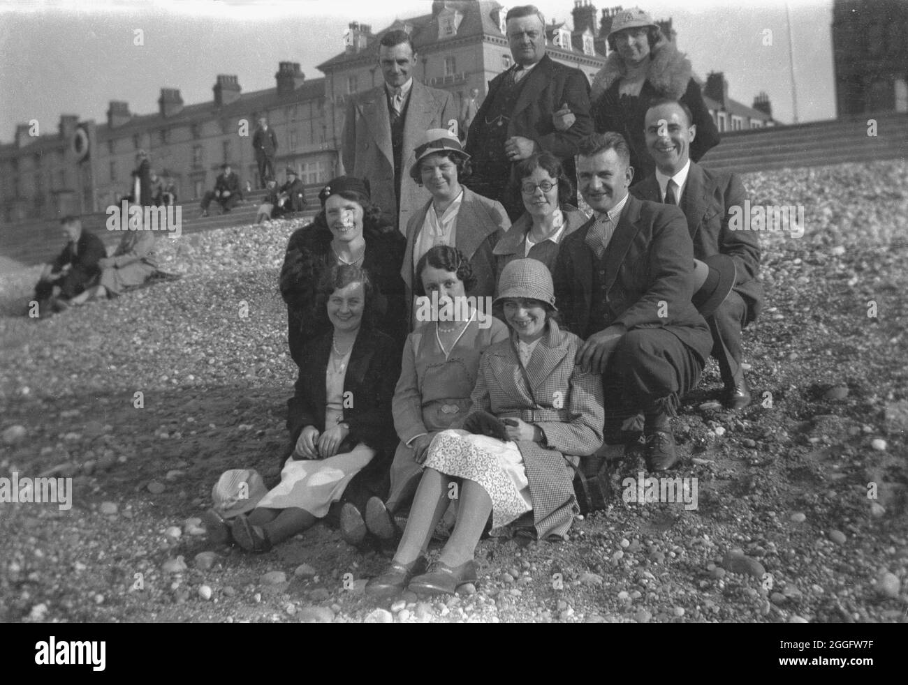 1930, histórico, dos familias se reúnen en una playa de piedras para una  foto de grupo. Llevan la ropa formal de la época, con los hombres en trajes  y corbatas y las