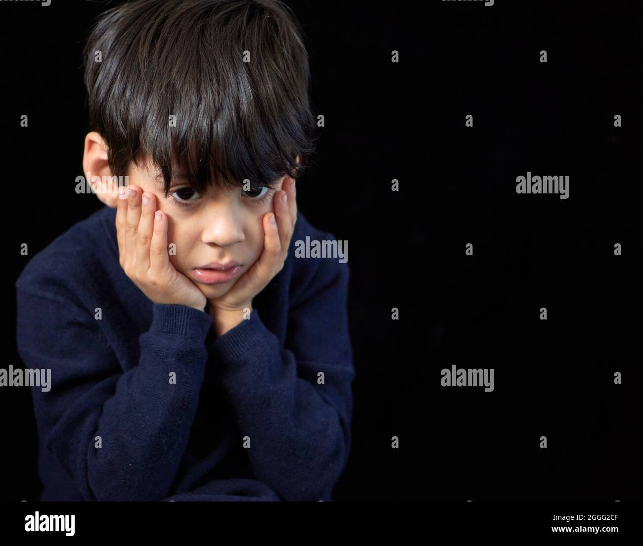 retrato de primer plano de un niño latino con una mirada fija en blanco preocupado y pensivo descansando su rostro en sus manos. fondo negro y copia Foto de stock